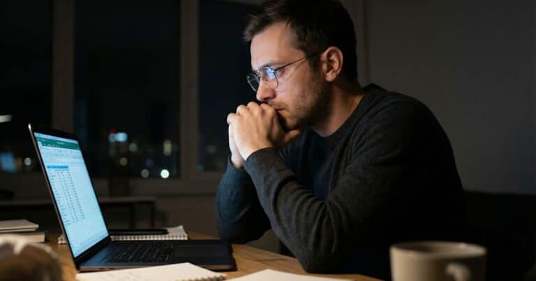 Bootstrapped founder reviewing spreadsheet at night in dim home office, reflecting on small business growth pressure